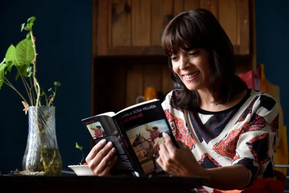 Camila Sosa sentada en interior, sonriendo mientras sostiene y lee un ejemplar de Las malas.