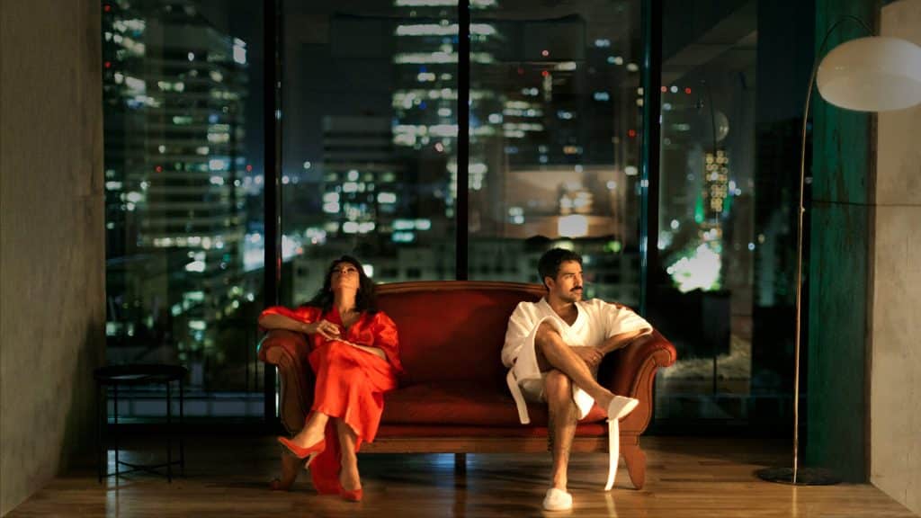 Camila Sosa Villada y el actor Alfonso Herrera comparten una escena en Tesis sobre una domesticación, sentados en un sofá frente a una vista nocturna de la ciudad.