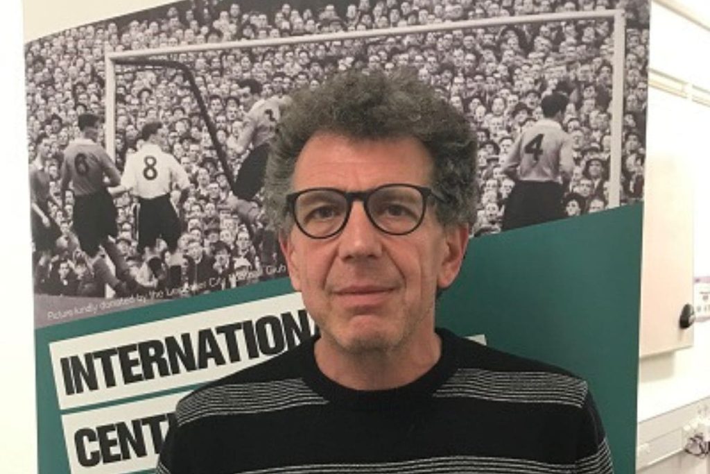 Retrato de David Goldblatt frente a una foto histórica de un partido.