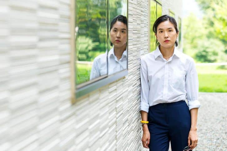 Retrato de Bora Chung junto a una pared de piedra, con camisa blanca y pantalón oscuro; su reflejo aparece en una ventana.