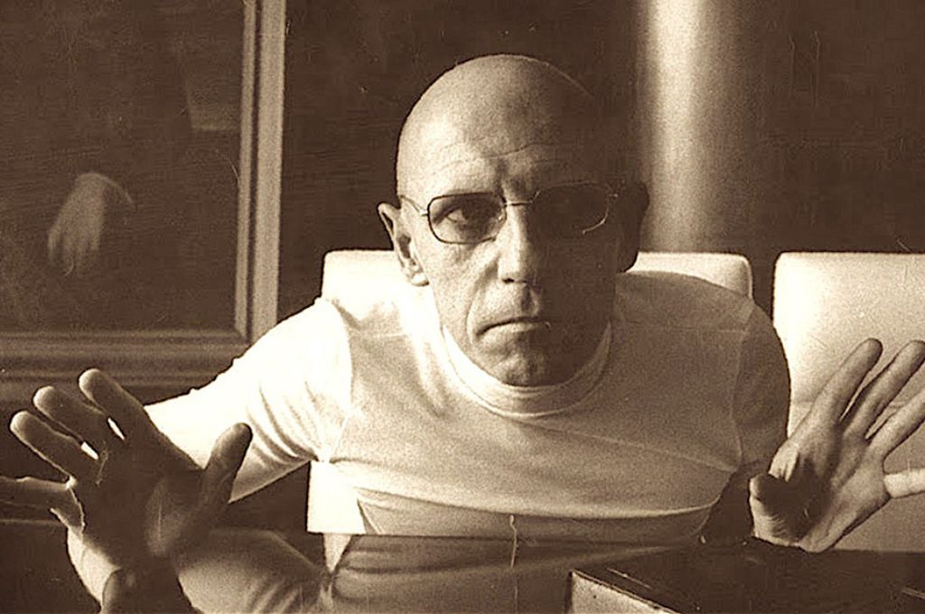 Michel Foucault en una imagen en primer plano, con las manos alzadas, gesto serio y lentes oscuros.