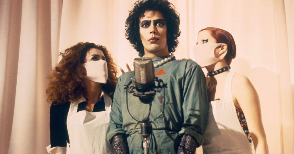Tim Curry como el Dr. Frank-N-Furter, flanqueado por dos mujeres con mascarillas blancas, frente a un micrófono vintage.