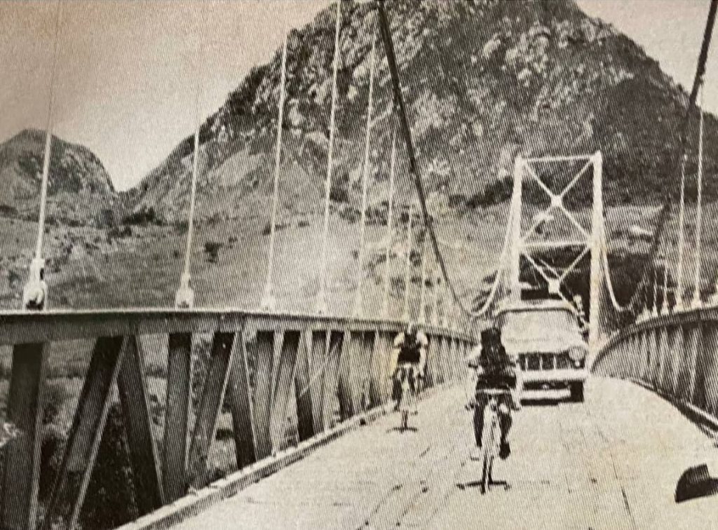 José Armando Alfaro cruza un puente colgante con una camioneta de apoyo detrás; montaña rocosa al fondo; foto en blanco y negro, década de 1950.