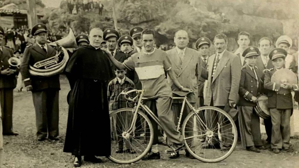 José Armando Alfaro con su bicicleta, rodeado por un sacerdote, músicos de banda y autoridades durante un acto público en los años cincuenta.
