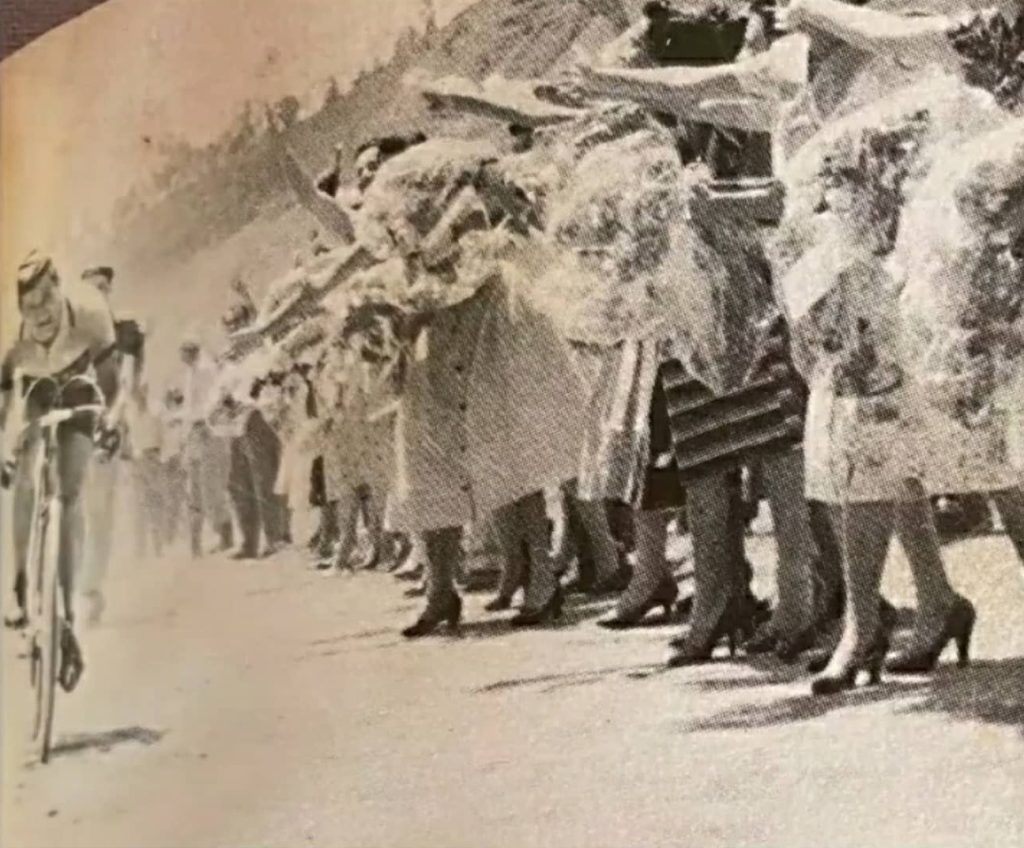 El ciclista avanza junto a una fila de mujeres que extienden ramos durante una etapa de la Vuelta a Colombia, años 50.