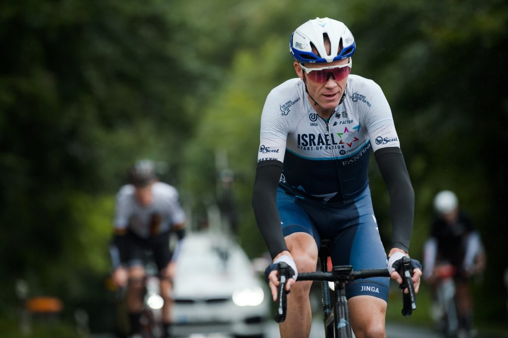 Primer plano de Chris Froome en el Israel Start-Up Nation, pedaleando bajo cielo nublado; casco blanco, gafas oscuras y maillot blanco-azul, con compañeros desenfocados al fondo.Equipo que es blanco del sportswashing en el ciclismo.