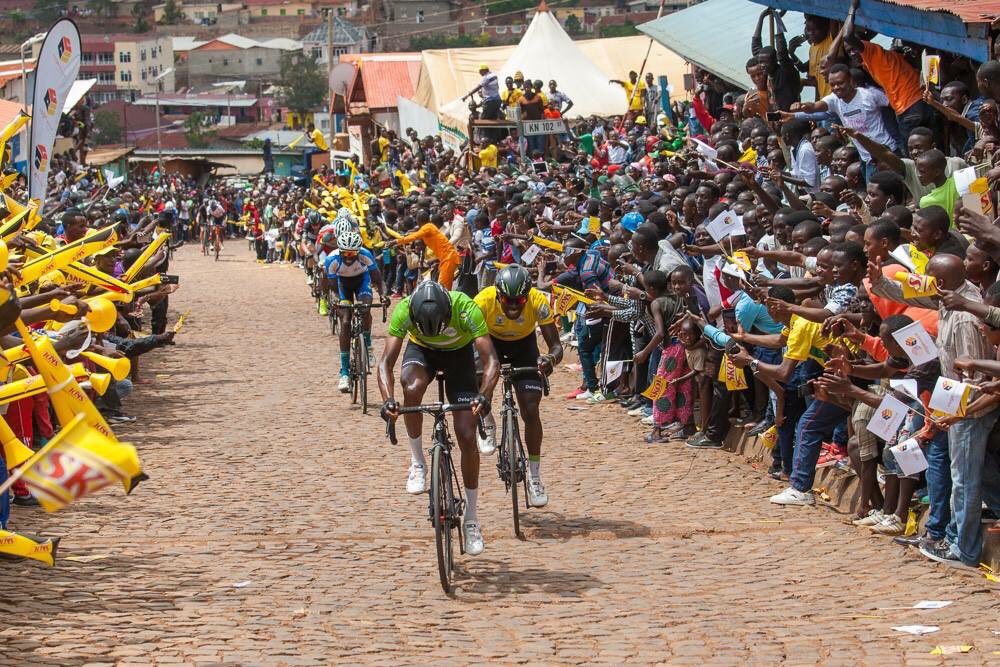 Multitud anima a dos ciclistas que coronan una subida empedrada durante el Tour de Ruanda; público a ambos lados agitando banderines y vuvuzelas. Este país es cuestionado por sportswashing en el ciclismo.