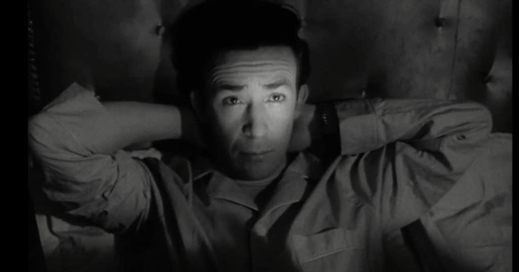 Hombre recostado en una cama, con las manos detrás de la cabeza, en una escena en blanco y negro de La frontera del sueño.