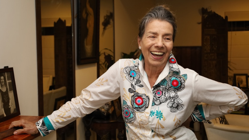 Bertha Quintero sonriendo, con el cabello recogido y una blusa blanca bordada con flores de colores, en el interior de una casa.