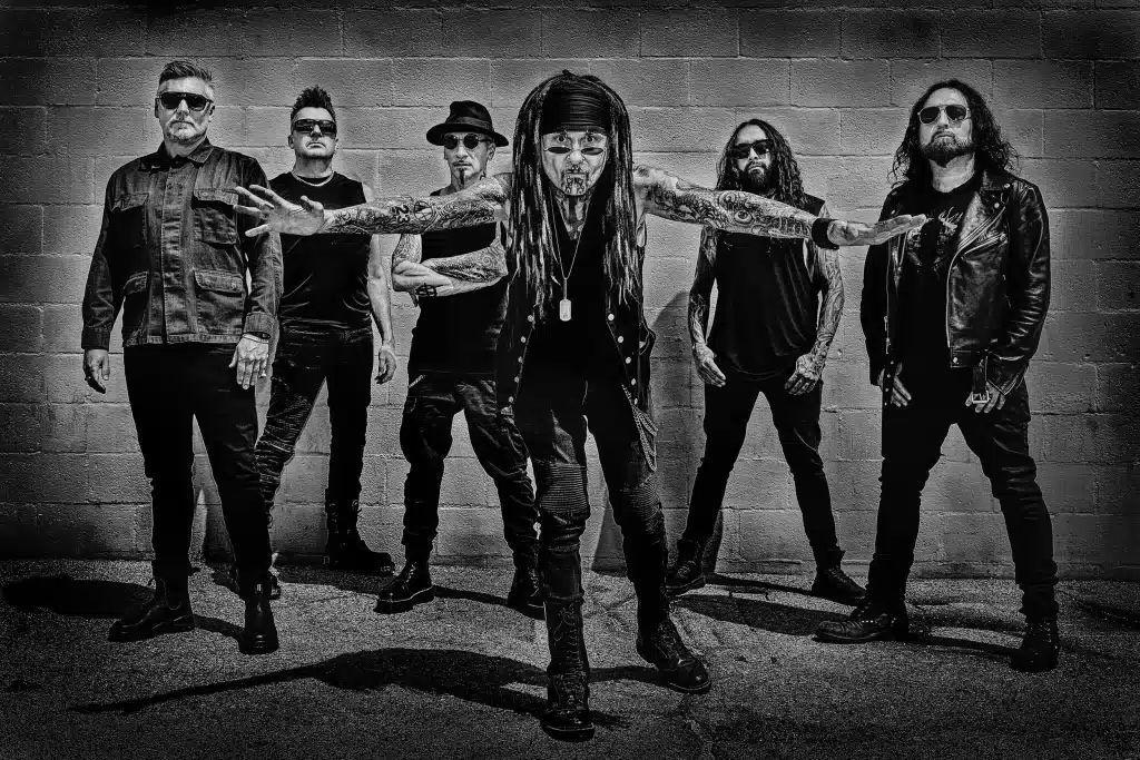Fotografía en blanco y negro de la banda Ministry: seis integrantes con ropa oscura y gafas de sol posan frente a un muro de bloques, y el del centro, de rastas, extiende los brazos tatuados